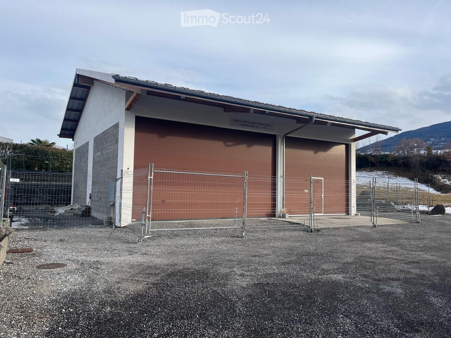 A single-story building with a large garage door, surrounded by a fenced area with gravel ground.