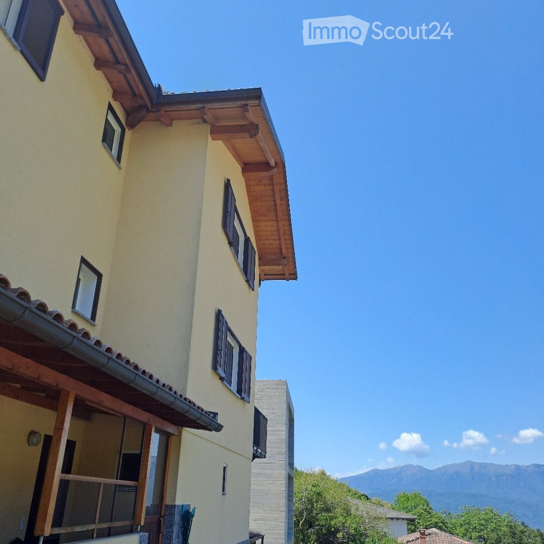 Two-story house with light yellow walls, wooden roofline, multiple windows with shutters, and a balcony on the first floor. The background shows a scenic mountain view.