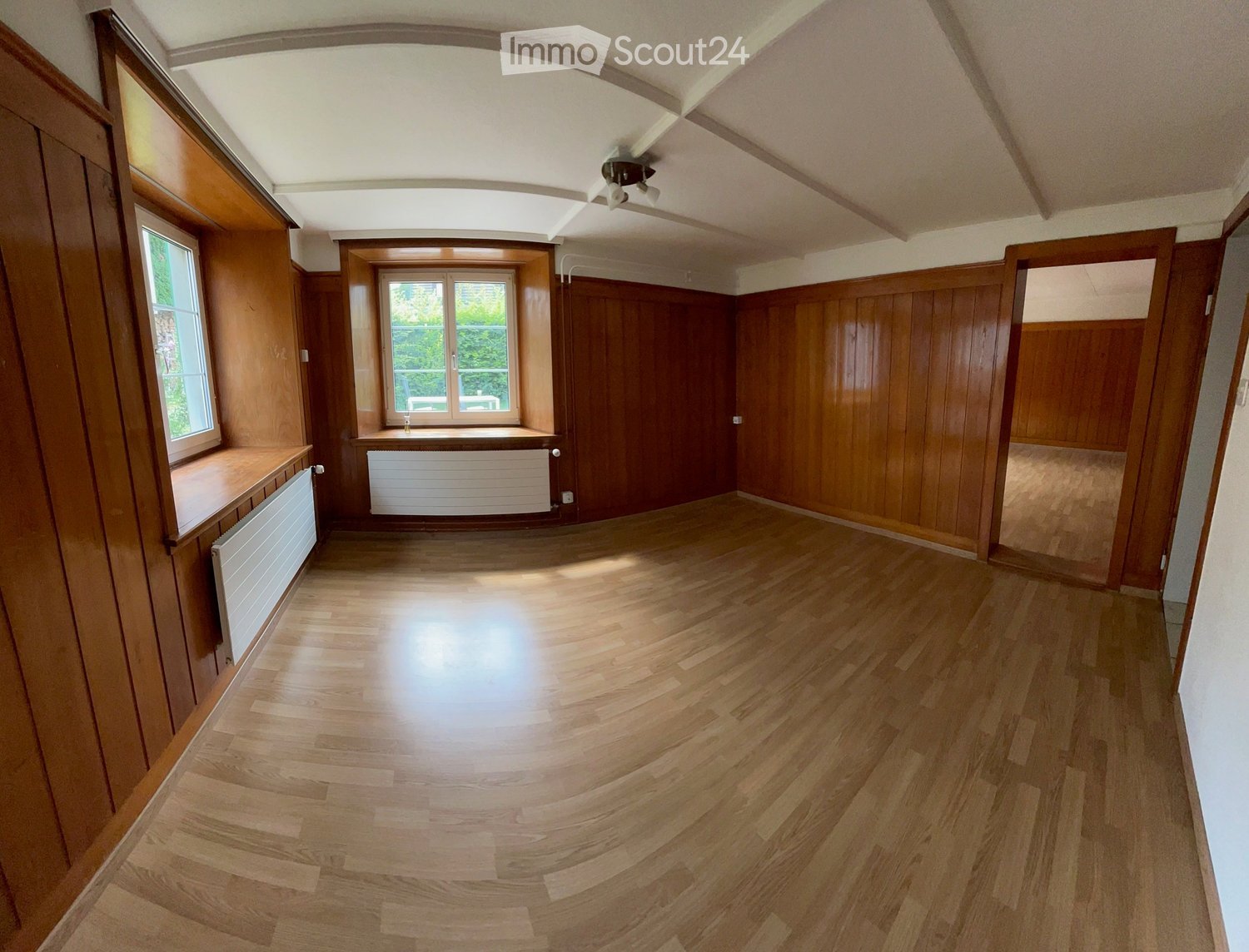 Empty room with wooden flooring, wooden panel walls, large windows, radiators, and a ceiling light fixture.