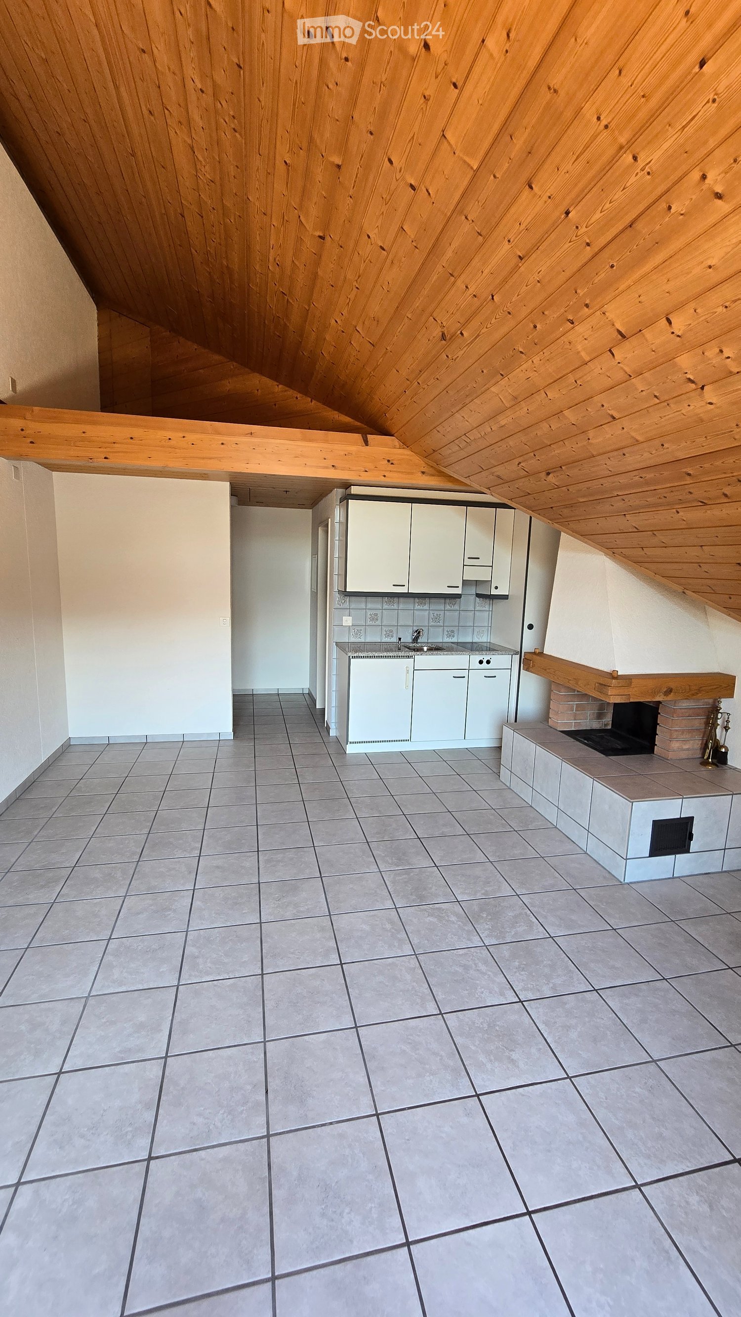Empty kitchen with white cabinets, a sink, and a fireplace, featuring a slanted wooden ceiling and tiled floor.
