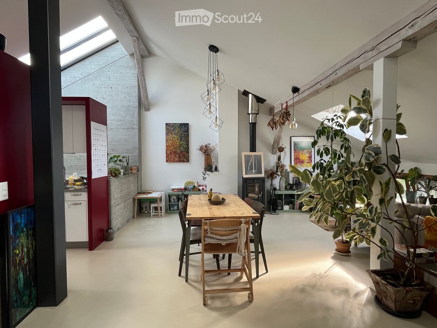 Bright, open living and dining area with exposed wooden beams, skylight, dining table with chairs, kitchen area with cabinets and counter, fireplace, potted plants, and artwork.