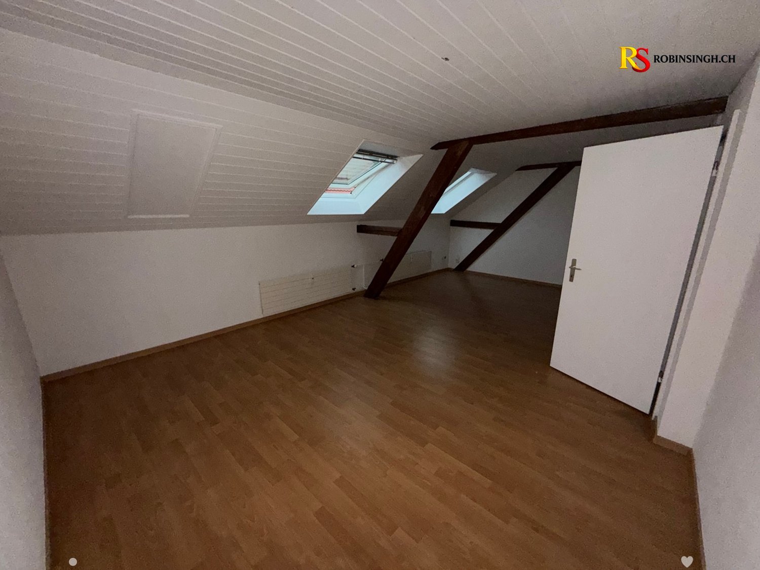 Empty attic room with wooden flooring, white walls, exposed wooden beams, and skylight windows.