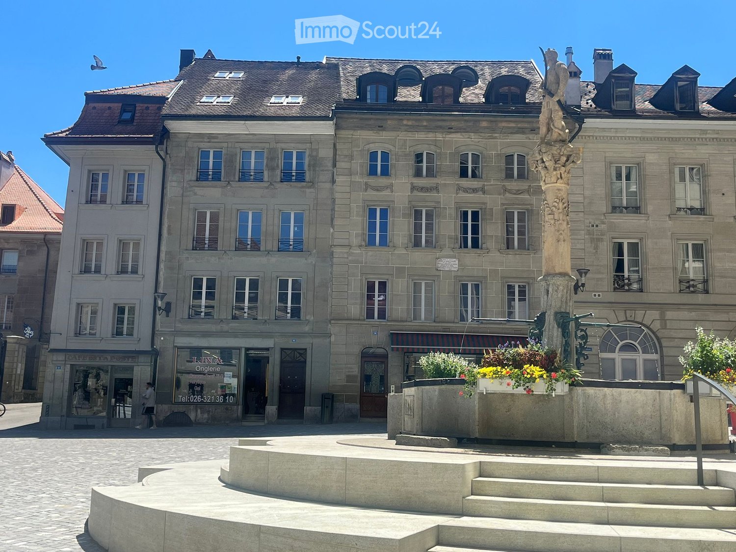 A large, multi-story building with a classic facade, numerous windows, and a decorative statue in the foreground. The ground floor features shops with signage, and the area is paved with cobblestones.