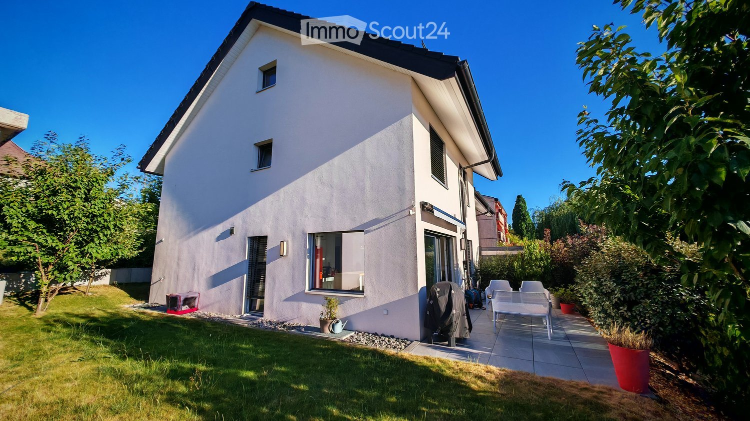 Modern two-story house with white exterior, large windows, and a well-maintained garden with patio seating.
