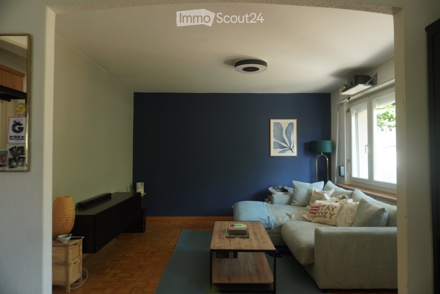 Modern living room with dark blue accent wall, light grey sectional sofa, wooden coffee table, side table, floor lamp, and decorative items.