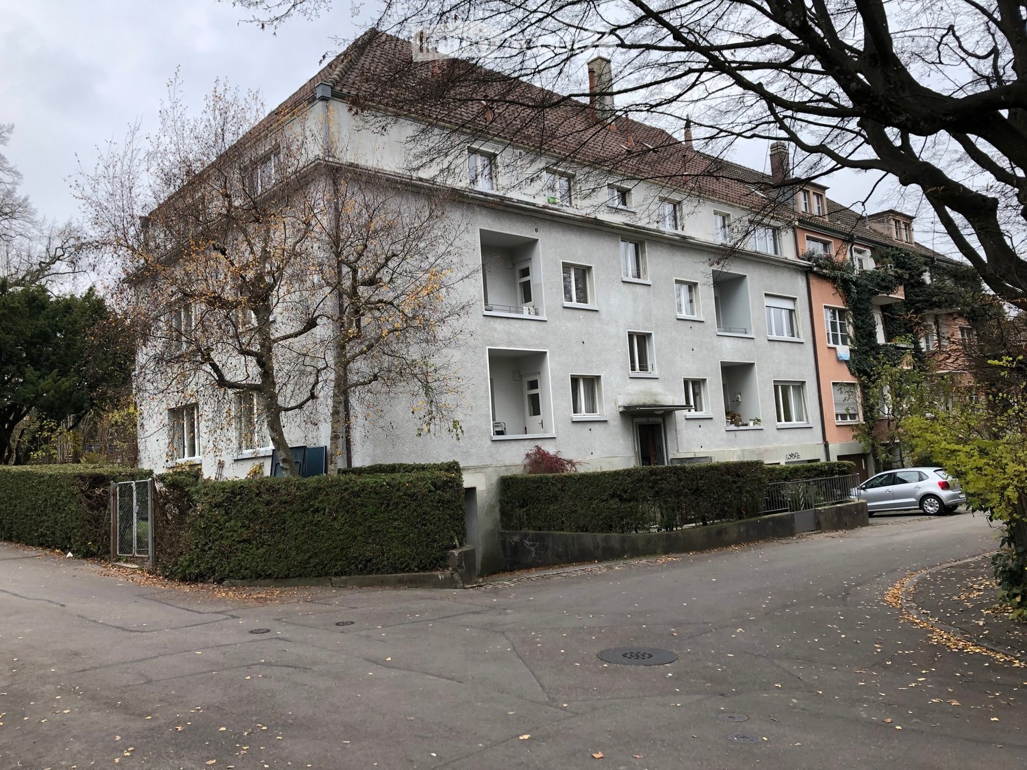 A multi-story residential building with white walls, multiple windows, and balconies, surrounded by trees and a parked car.