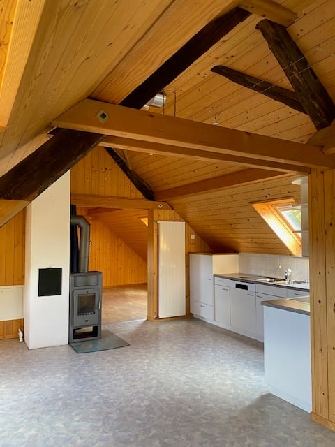 Open-plan attic space with exposed wooden beams, kitchen area with white cabinets, sink, and faucet, and a fireplace with a wooden floor.