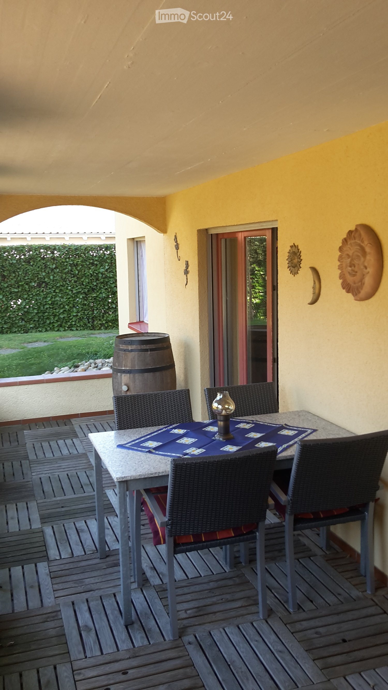 Wooden deck with a round table, four wicker chairs, and a small lamp in the center. The table is covered with a blue placemat and has playing cards on it. There is a large barrel in the background and a view of a garden through an open doorway.