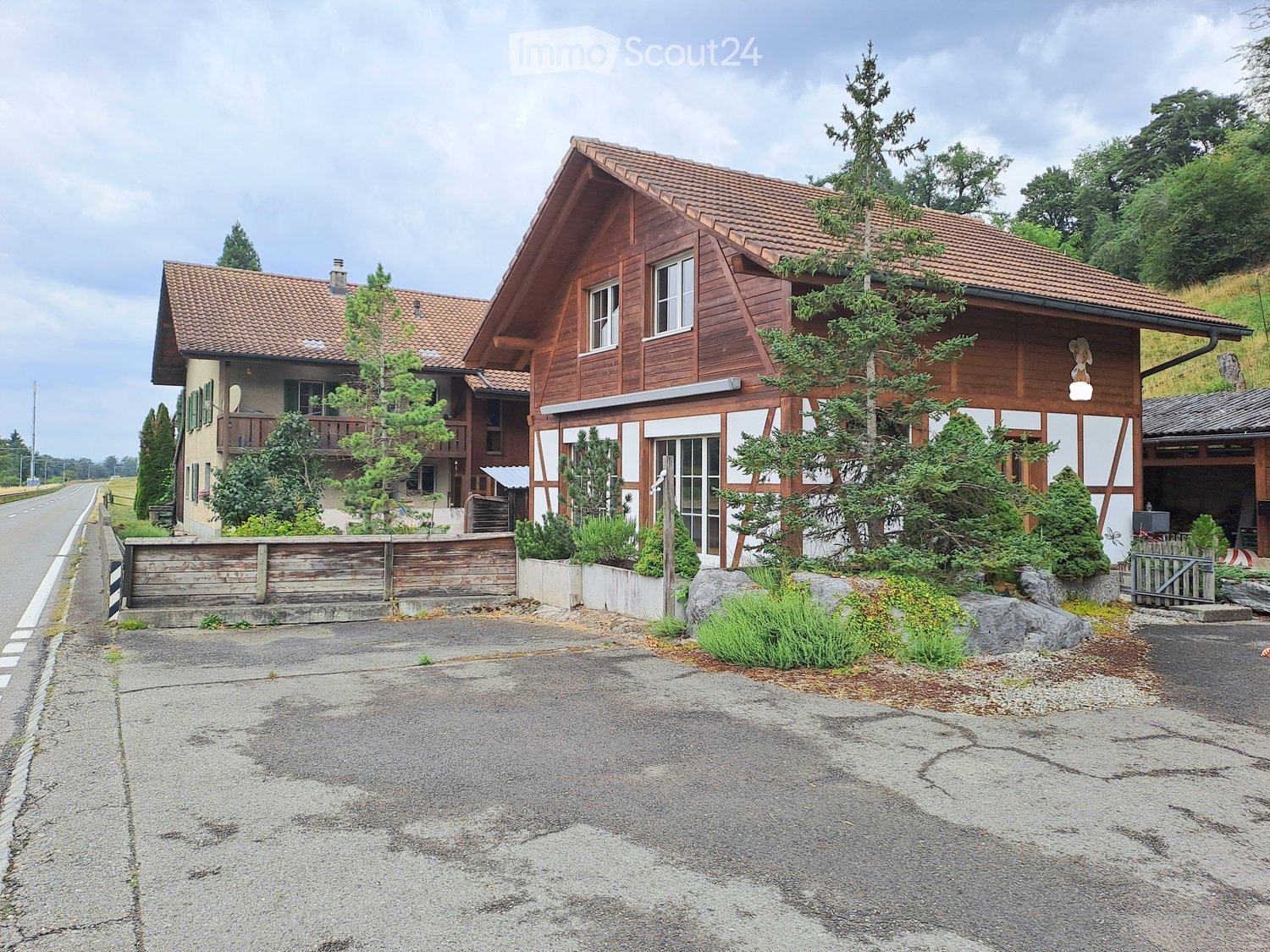 Two-story wooden house with red-tiled roof, multiple windows, and a small balcony on the left side. The house is surrounded by greenery and a wooden fence.