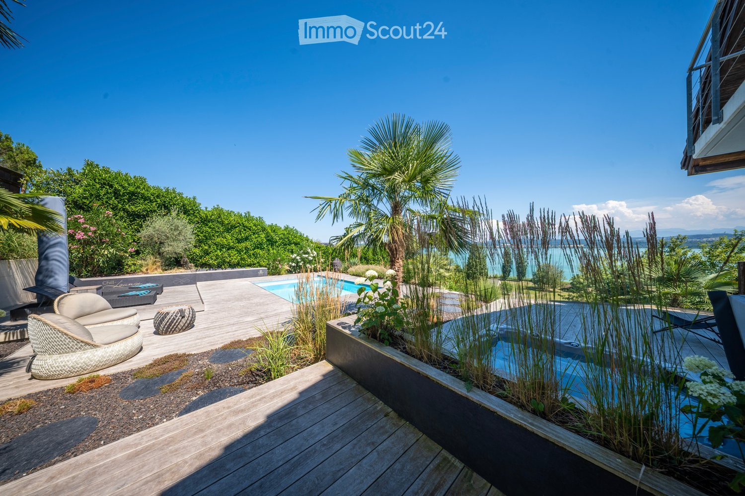 A modern outdoor area with a swimming pool, lounge chairs, a small seating area, and lush greenery, including palm trees and tall grasses, overlooking a scenic lake view.