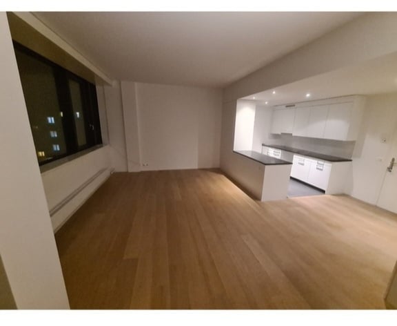 Bright, open-plan living and dining area with light wood flooring, large window, and a modern white kitchen with cabinets and countertop.