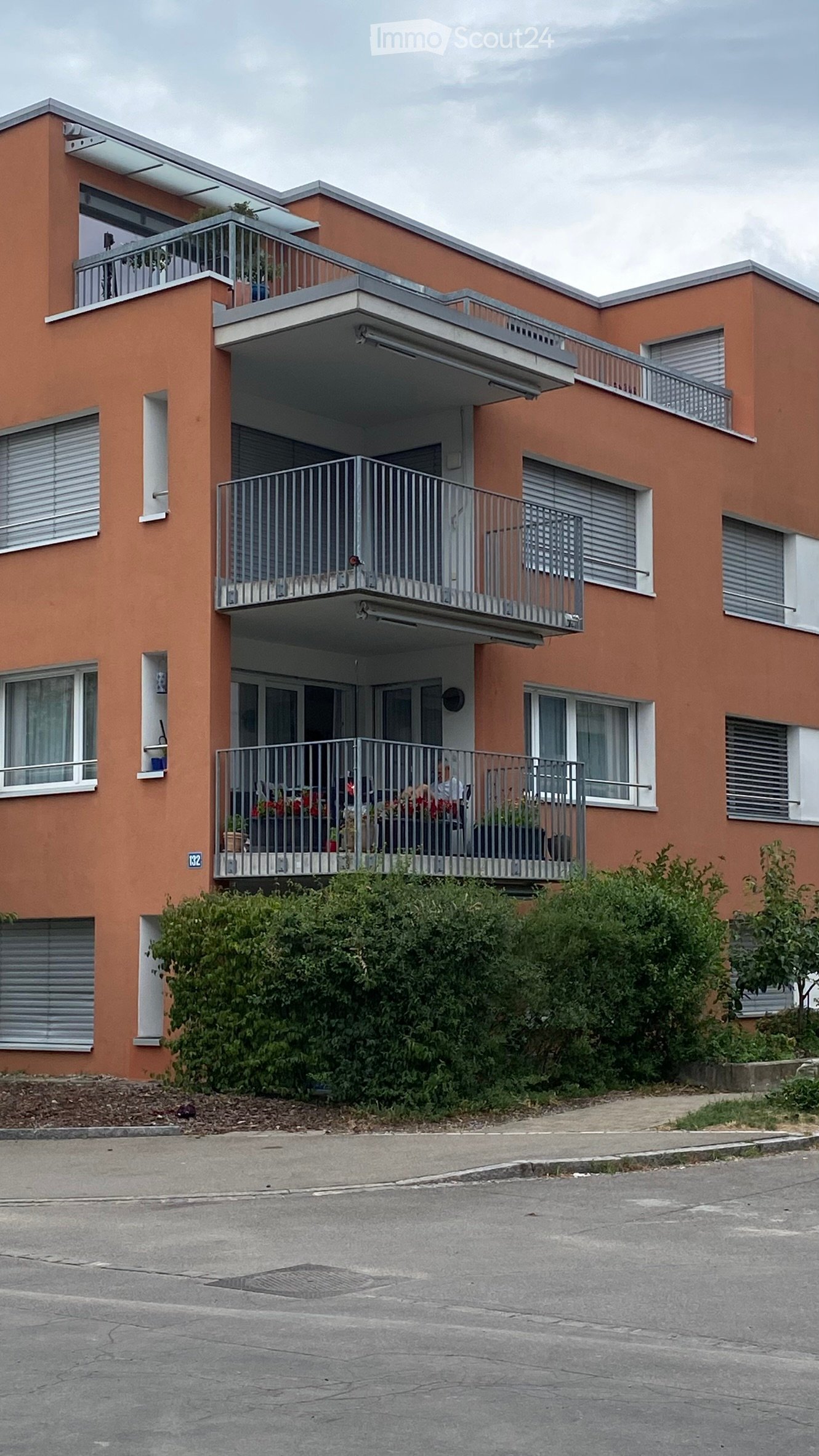 Modern three-story apartment building with orange facade, multiple balconies, and greenery in front.