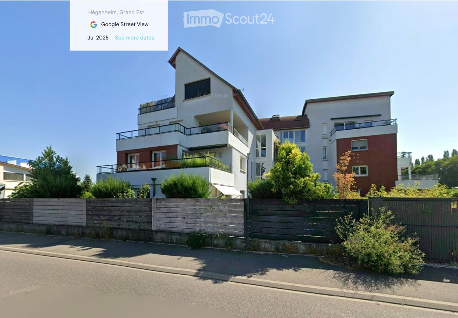 Modern multi-story residential building with balconies, surrounded by greenery and a wooden fence.