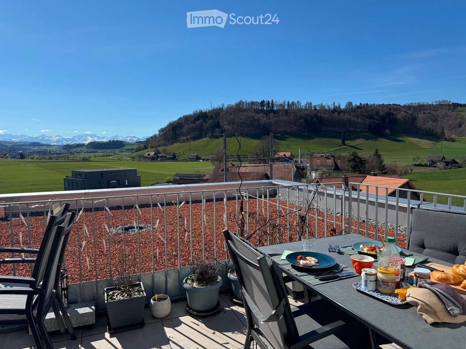 Balcony with a scenic view of green fields and mountains, equipped with a table, chairs, and a small plant.