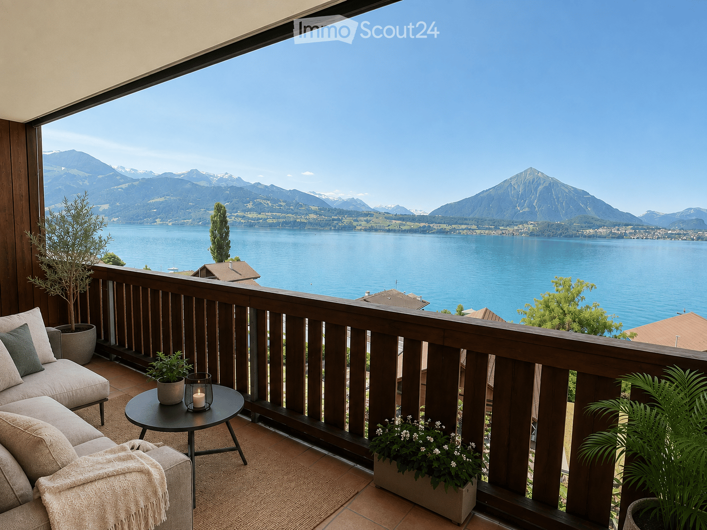 Balcony with wooden railing, overlooking a lake and mountains, furnished with a sofa, small table, candle, and potted plants.