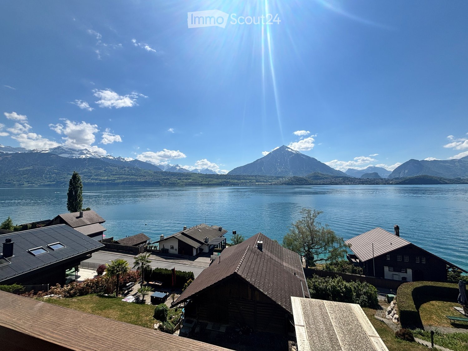 View of a serene lake surrounded by mountains, with several houses and a well-maintained garden in the foreground.