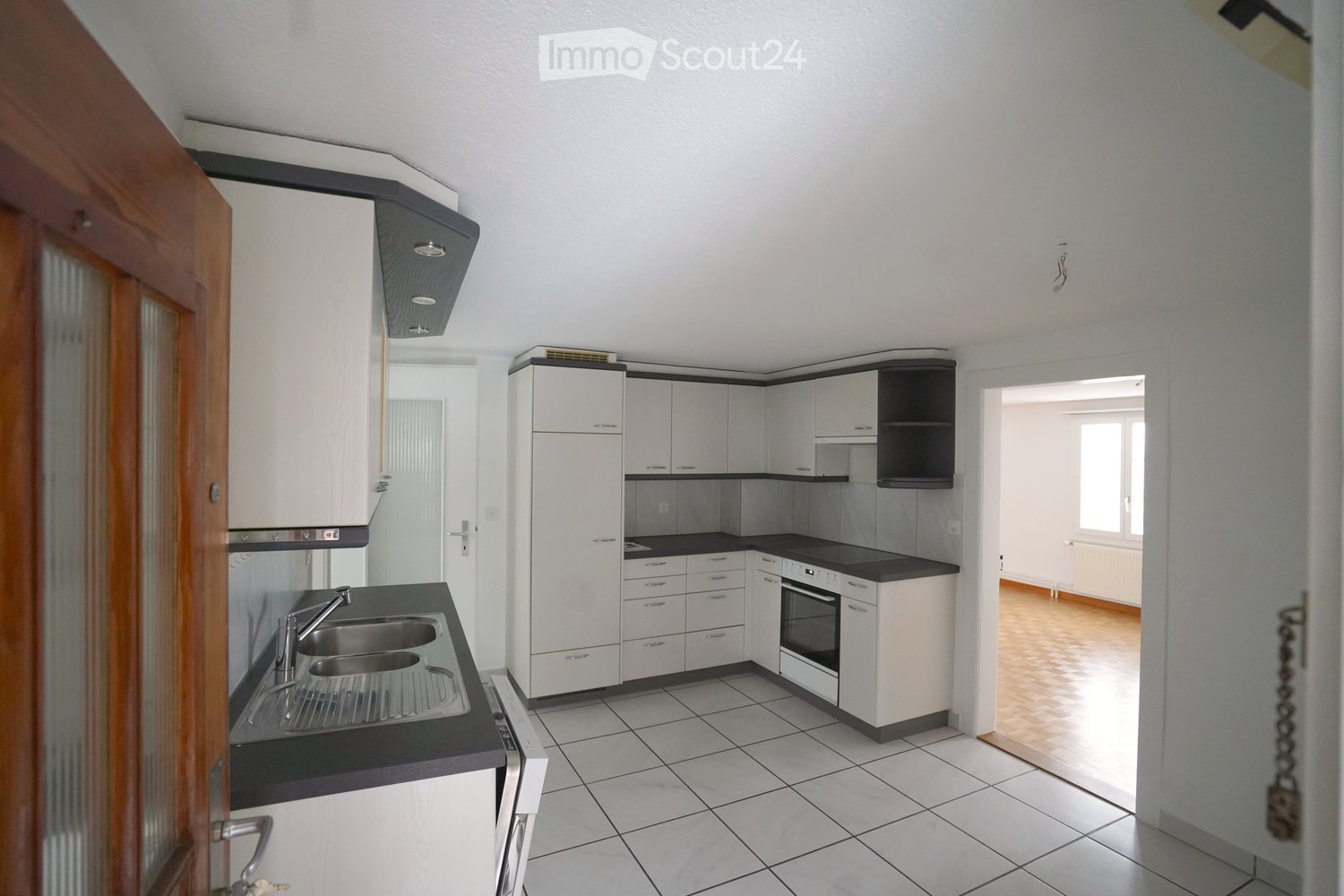 Modern kitchen with white cabinets, black countertops, stainless steel sink, and built-in oven. The space features tiled flooring and a wooden door leading to another room.