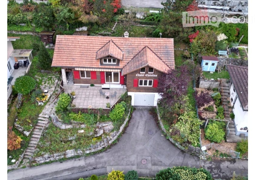 A two-story house with a red-tiled roof and red shutters, surrounded by a well-maintained garden and driveway. The house features a garage and a small patio area with outdoor furniture.