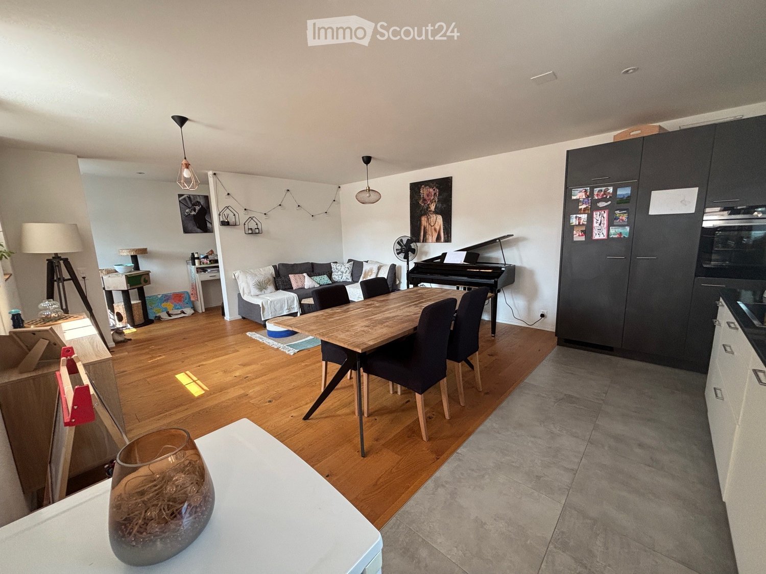 Open-plan living and dining area with wooden flooring, a sofa, dining table with chairs, piano, and various decorative items.