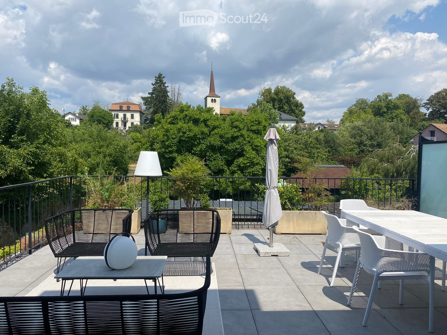 Balcony with outdoor furniture including a sofa, table, chairs, and an umbrella, overlooking a scenic view of trees and a church spire.
