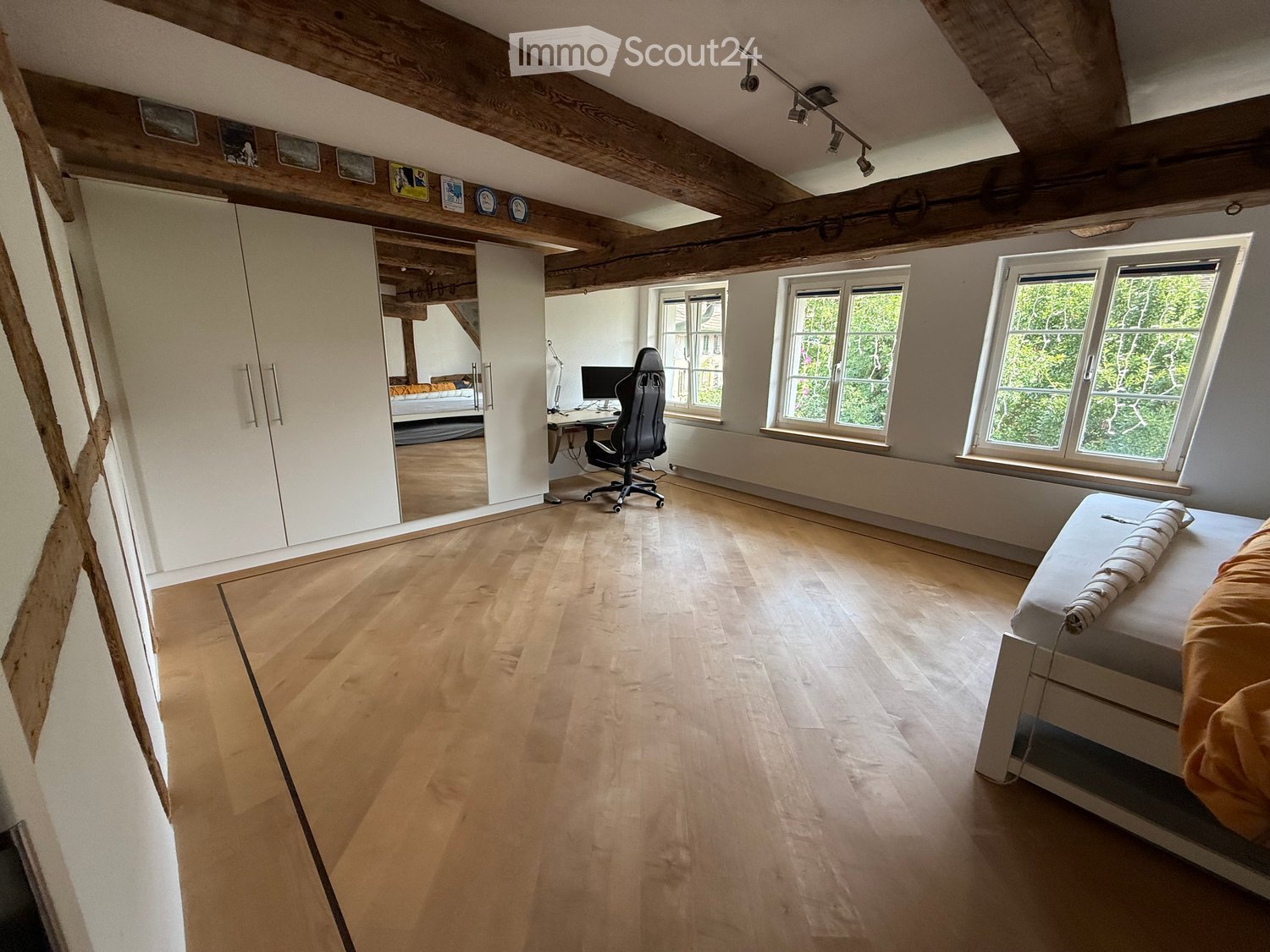 Bright bedroom with wooden floors, exposed wooden beams, large windows, a bed with orange bedding, a desk with a computer, and a wardrobe.