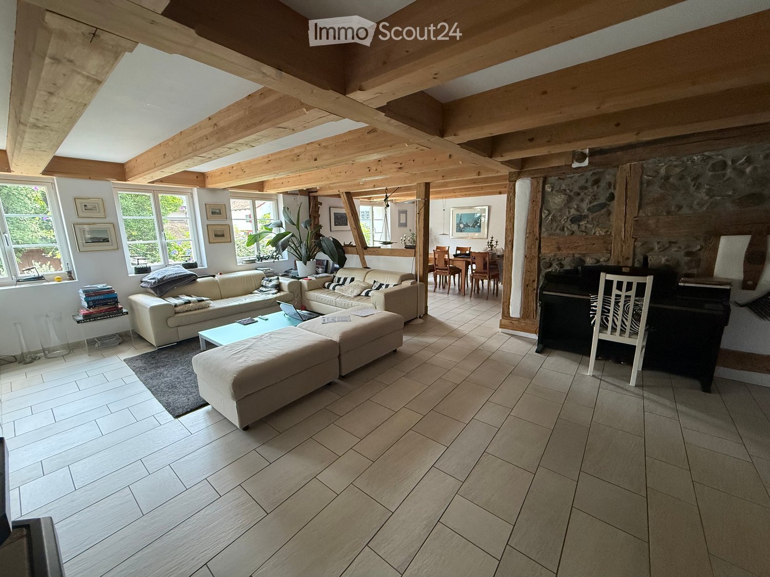 Bright living room with exposed wooden beams, large windows, cream-colored sofa, coffee table, ottoman, dining area with chairs and table, piano, and decorative items.