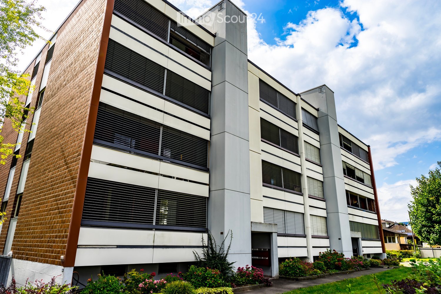 Modern multi-story building with a combination of brick and concrete facade, large windows with blinds, and a landscaped garden area in front.