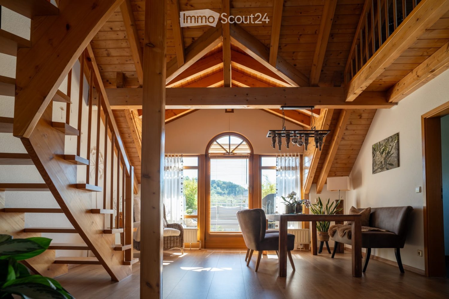 Bright living room with wooden beams, large windows, a staircase, a sofa, a chair, a small table, and a hanging chandelier.