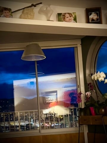 A cozy living space with large windows offering a city view, a floor lamp, a small table with a flower pot, and decorative items on the shelf above the window.