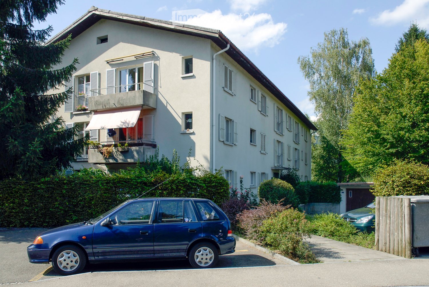 A two-story house with white exterior walls, multiple windows, and a balcony on the upper floor. The house is surrounded by greenery and has a driveway with a parked car.