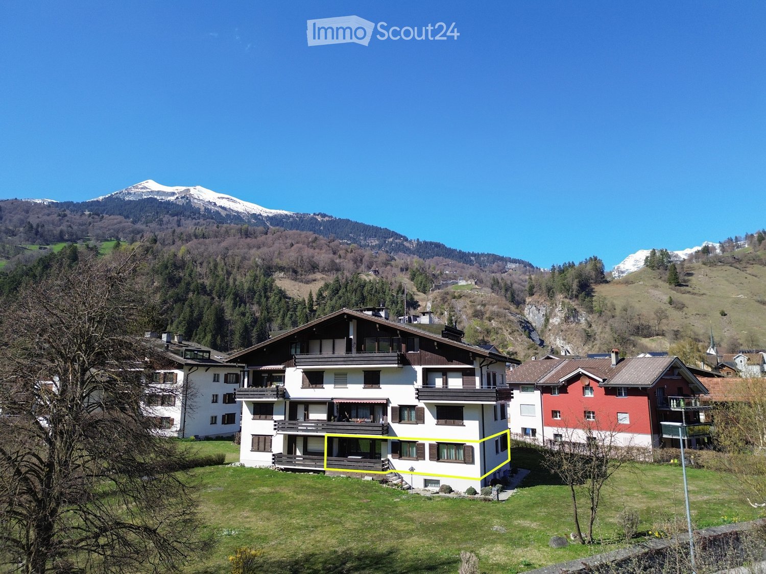 A multi-story house with white walls, dark brown trim, and multiple balconies, situated in a scenic valley with snow-capped mountains in the background.