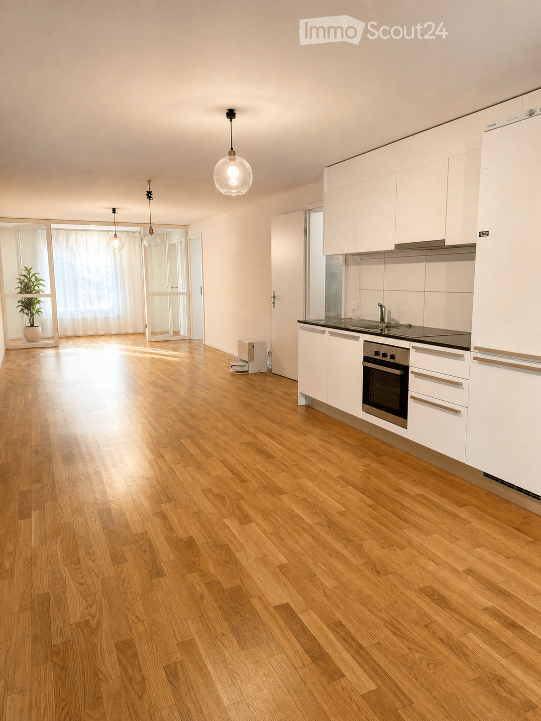 Modern kitchen with white cabinets, wooden flooring, and pendant lights. Features a refrigerator, oven, and sink.
