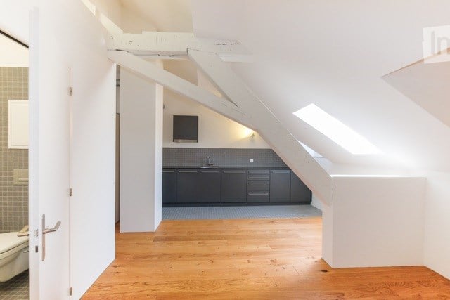 Loft-style kitchen area with grey cabinets, a sink, and a small screen mounted on the wall. The space features wooden flooring and exposed white beams.