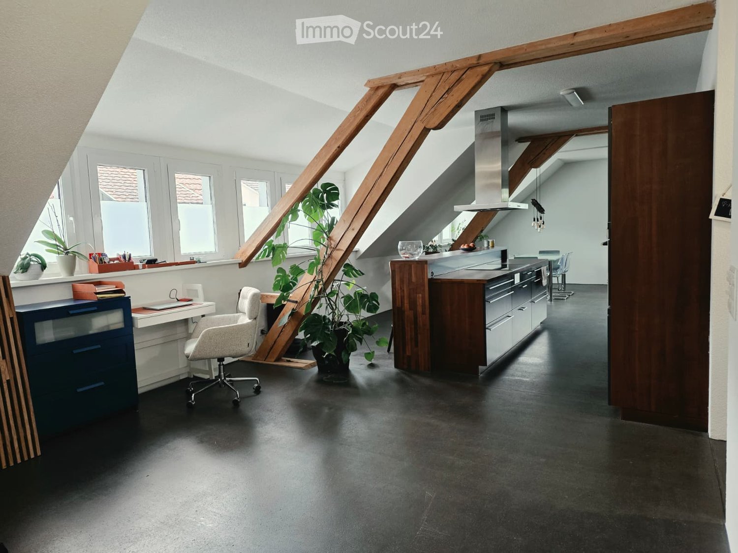 Open-plan attic space with a modern kitchen and a workspace area. Features include a grey office chair, desk with laptop, blue cabinet, large potted plant, and kitchen appliances like an oven, stove, and fridge.