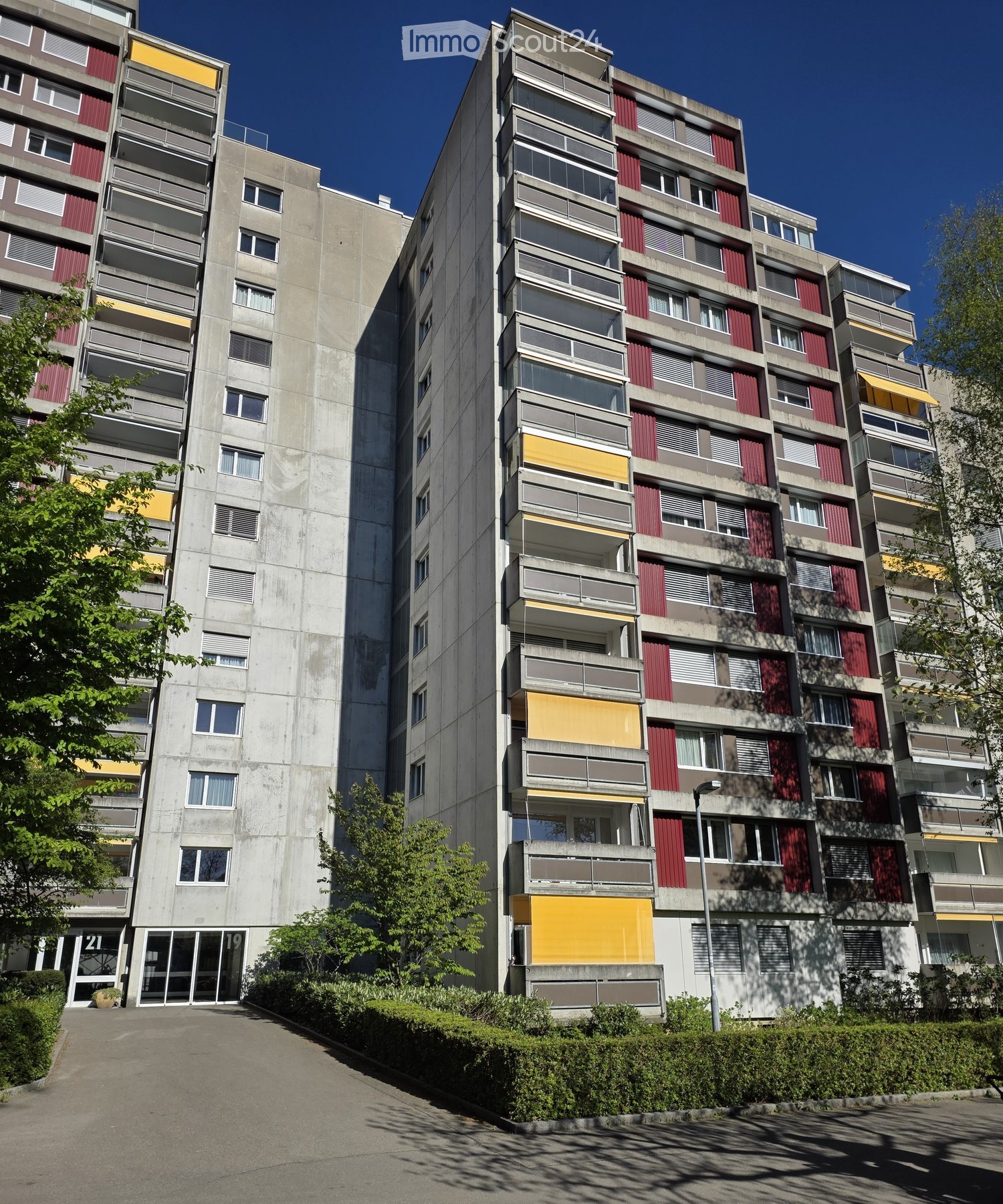 Multi-story residential building with concrete facade, red and yellow accents, multiple windows, and balconies.