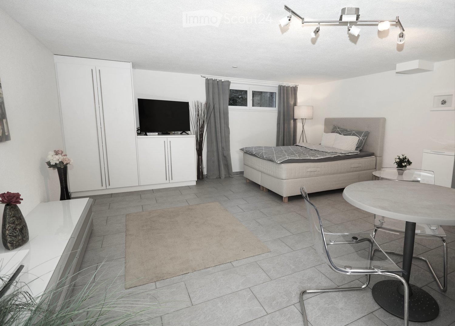 Modern bedroom with a large bed, bedside table, TV, wardrobe, and a small dining area with a table and chairs.