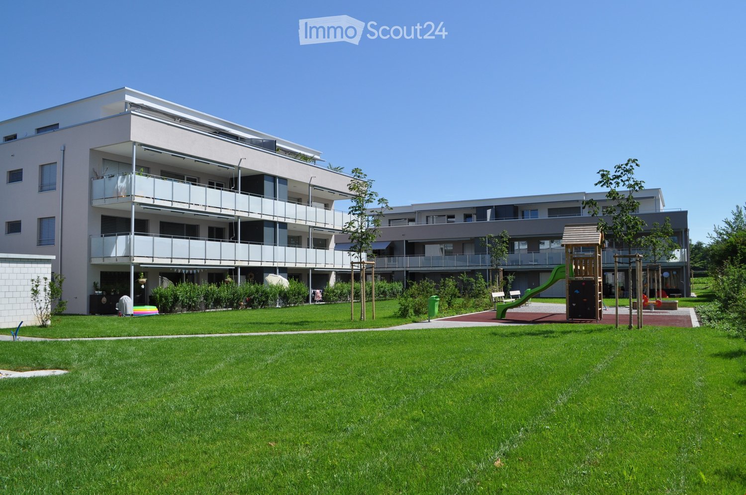 White apartment building with three floors, balconies, playground, grass lawn, plants, trees, and green slide.