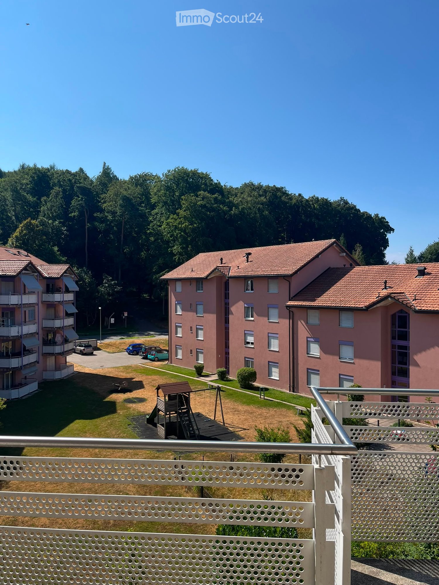 View from a balcony overlooking a residential complex with pink buildings, a playground, and a parking area.