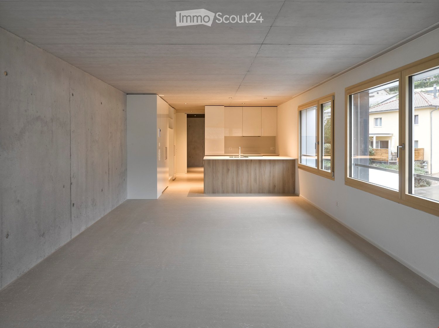 Open, minimalist space with concrete walls and floor, large windows, and a small kitchen area with light-colored cabinets and a wooden countertop.