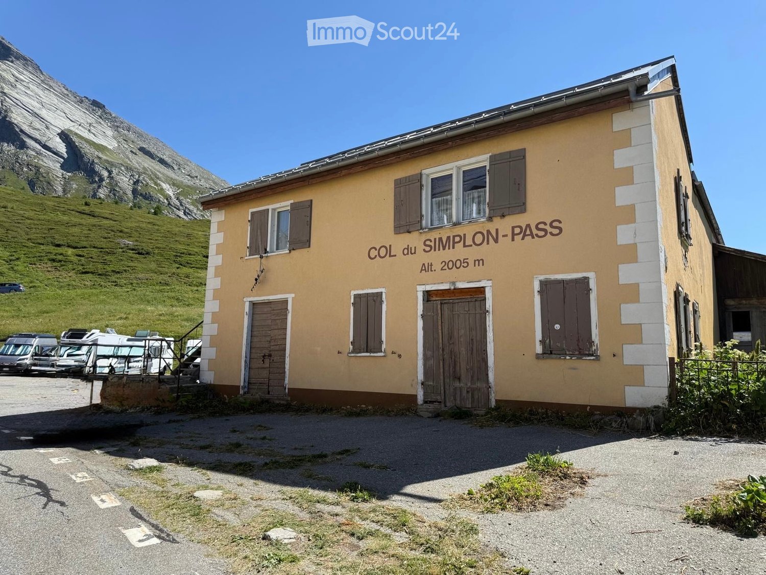 A small, yellow building with brown shutters and a wooden door, labeled "COL du SIMPLON-PASS" with an altitude of 2005 m. It is situated near a parking area with several vehicles and a mountainous backdrop.