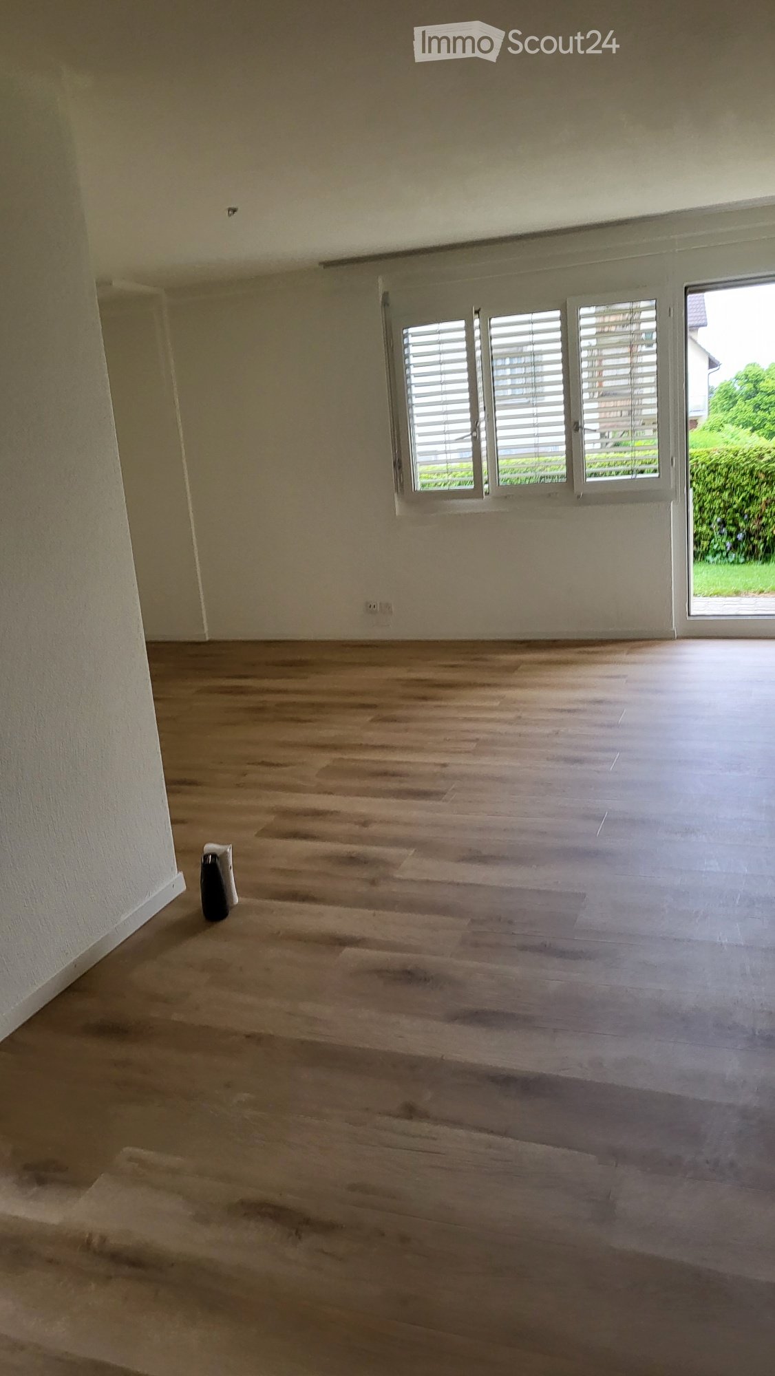 Empty room with wooden flooring, white walls, and large windows with blinds. A small black object is placed near the wall.