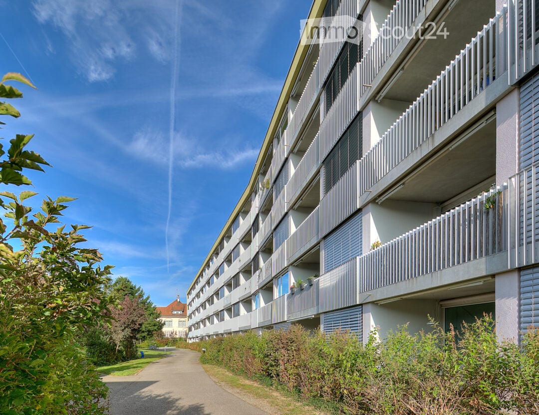 Modern multi-story apartment building with balconies, surrounded by greenery and a clear blue sky.