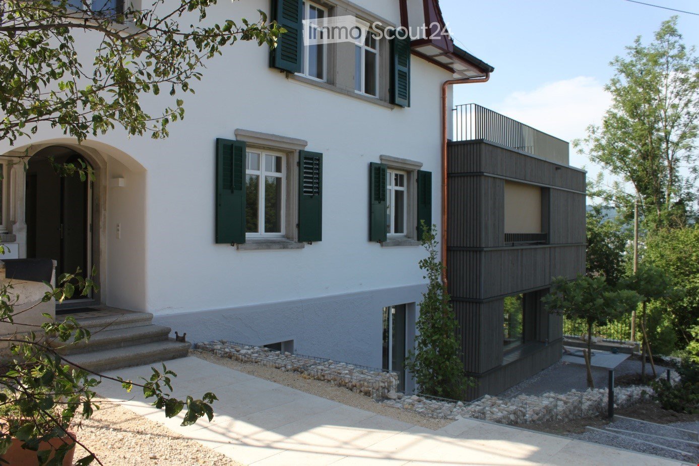 Two-story house with white exterior, green shutters, and a modern balcony on the upper level. The entrance features a small staircase leading to a dark door, and the ground floor includes a raised section with a stone border.