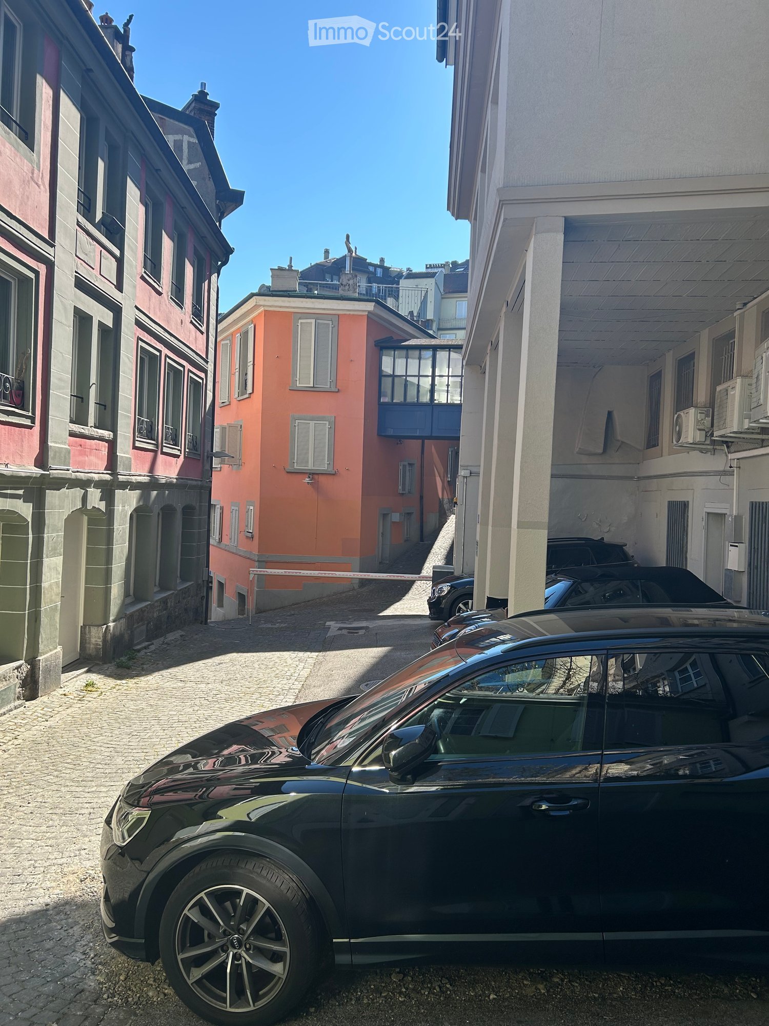 A narrow urban street lined with multi-story buildings, featuring a mix of architectural styles and parked cars.