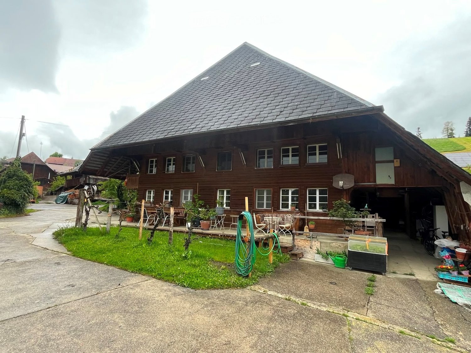 Traditional wooden house with a steep roof, multiple windows, and a spacious outdoor area with chairs, tables, and greenery.