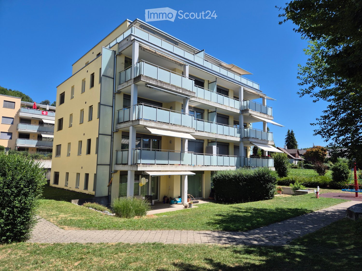 Modern multi-story residential building with balconies, surrounded by a well-maintained lawn and garden area.