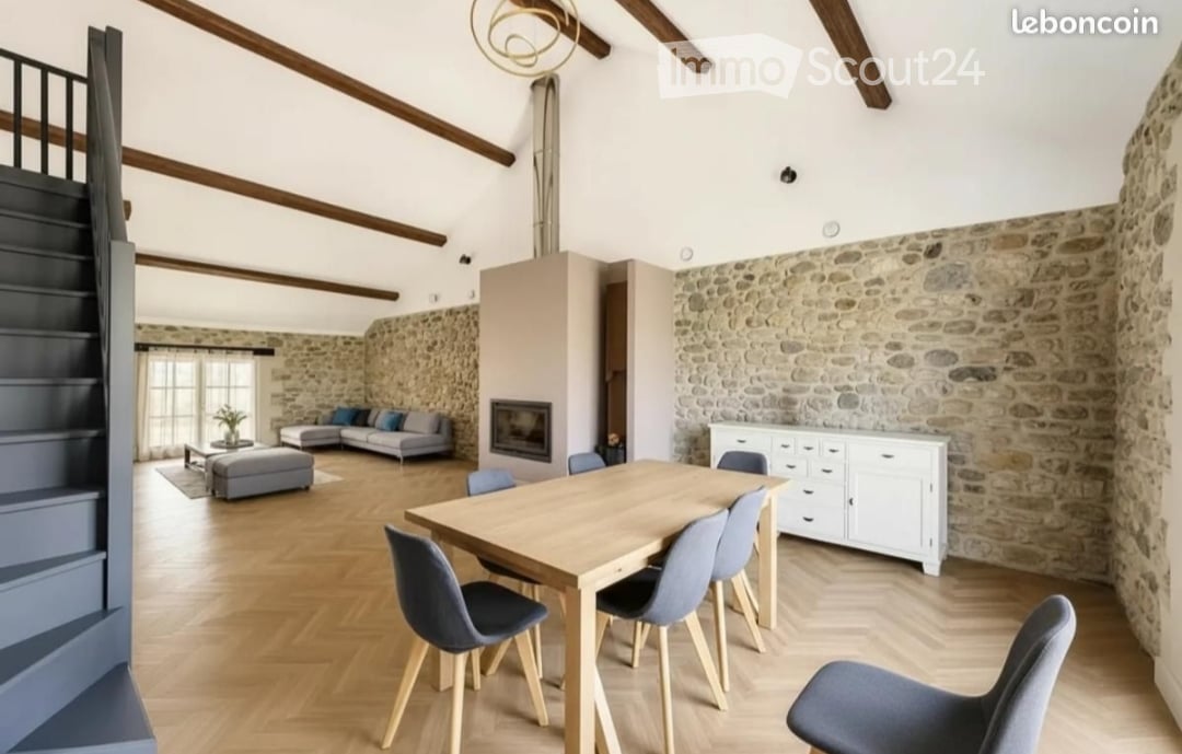 Open-plan living and dining area with exposed wooden beams, stone walls, fireplace, grey sofa, coffee table, ottoman, dining table with chairs, and white cabinet.