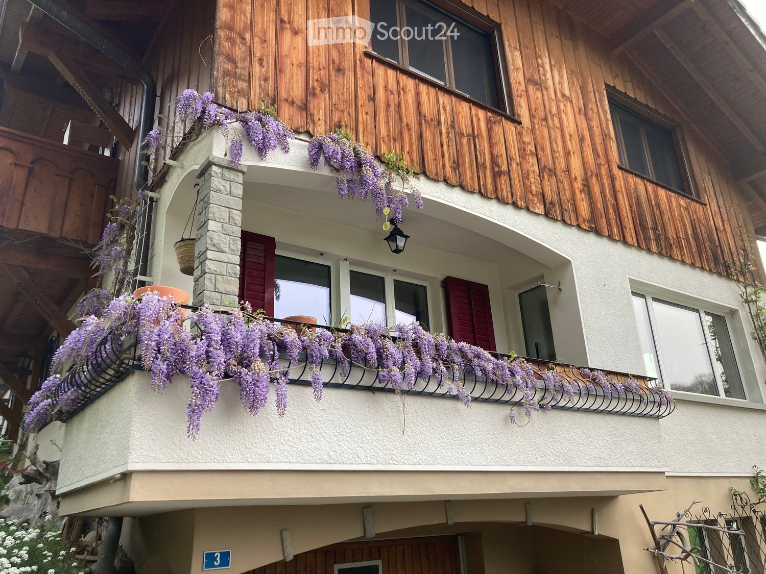 Balcony with purple wisteria flowers, wooden shutters, and a hanging basket. The building has a mix of wooden and plaster finishes.