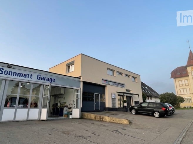 A modern, single-story building with a garage entrance labeled "Sonnmatt Garage." The building has a flat roof, large windows, and a clean, contemporary design. A black SUV is parked in front of the garage.