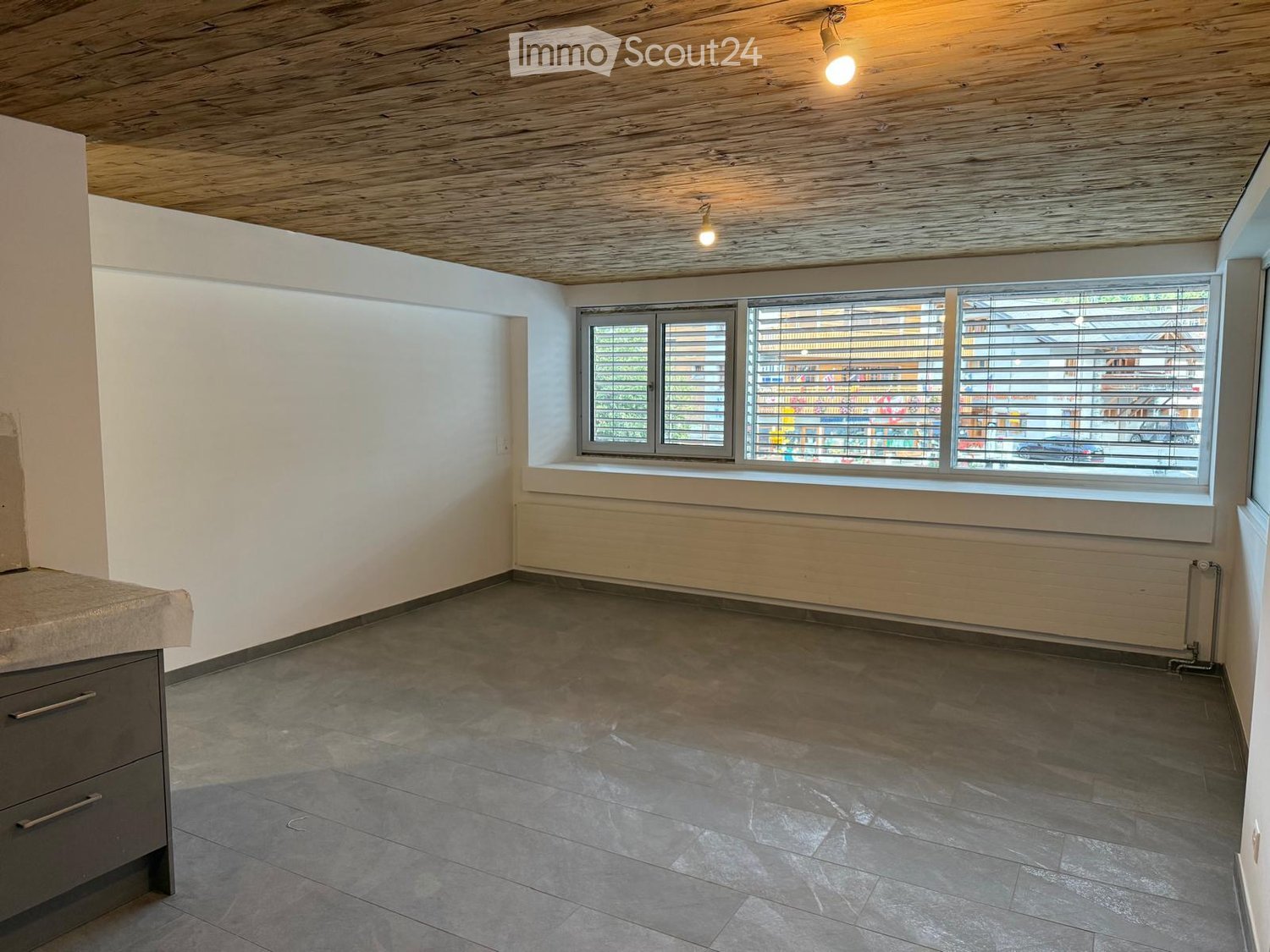 Empty room with wooden ceiling, tiled floor, large window with blinds, radiator, and a small cabinet with drawers.